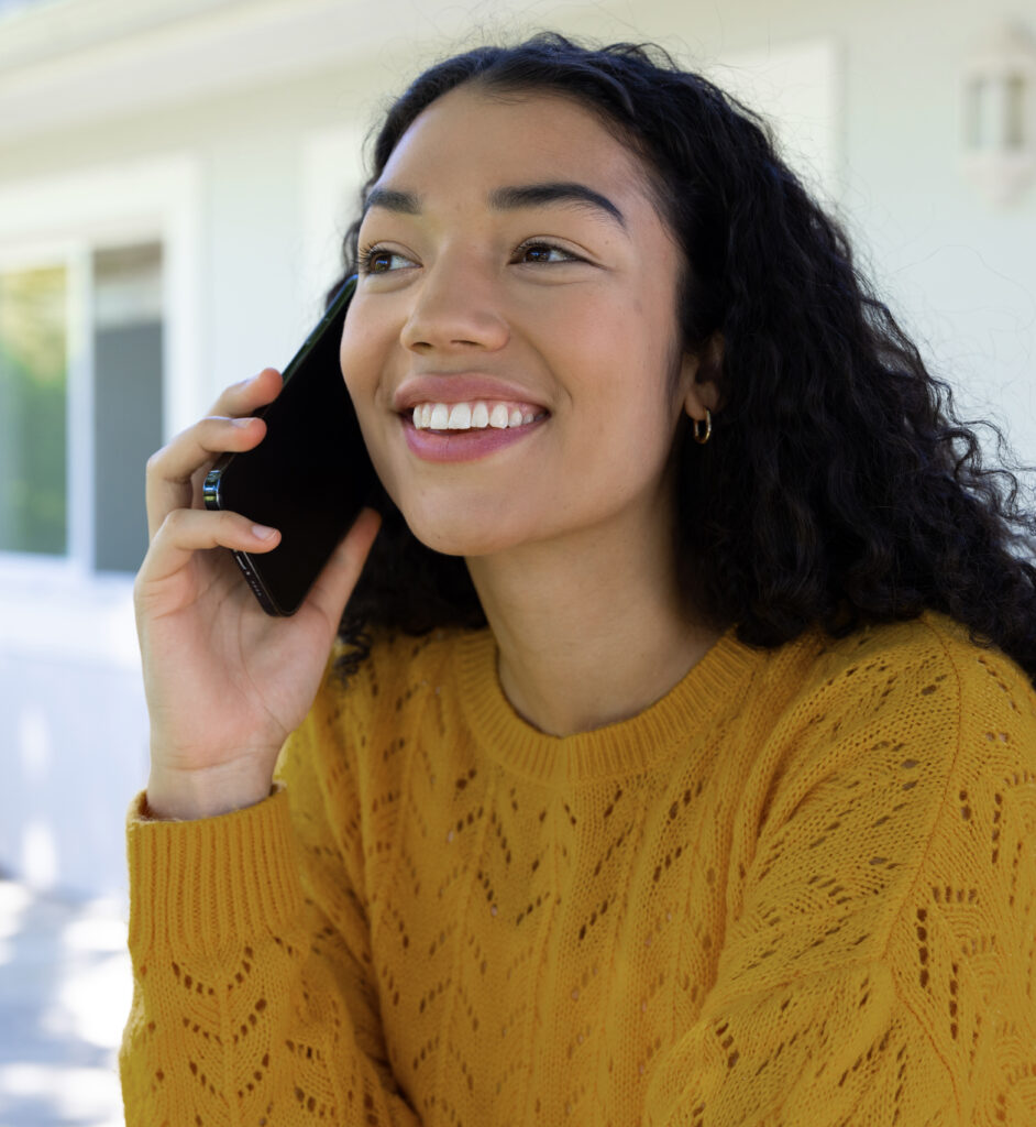 Happy biracial woman in yellow sweater talking on smartphone on sunny terrace. Lifestyle, communication and domestic life, unaltered.